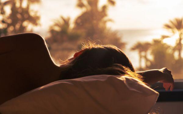 Silhouette of woman lying on the bed and sleeping on pillow in bedroom opposite panoramic window with view on palm tree sea beach at sunset. Back view.